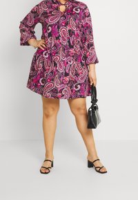 Vestido de estampado paisley rosa con escote en forma de lágrima y mangas three-quarter, combinado con sandalias negras de tacón y un pequeño bolso negro.