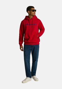 Czerwony bawełniany hoodie z niebieskim napisem "diverse", przednia kieszeń i sznurek. Noszony z niebieskimi dżinsami i białymi trampkami, uzupełniony czarnymi okularami przeciwsłonecznymi.