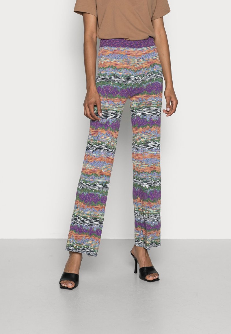 Personne portant un pantalon en maille rayé multicolore avec une jambe évasée et des sandales à talons ouvertes noires sur fond blanc.