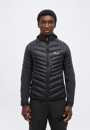 Młody mężczyzna w czarnej pikowanej kurtce z kapturem z pomarańczowym suwakiem i czarnych spodniach, stojący na tle jednolitego białego tła.