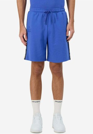 Homme portant un short de sport bleu avec une bande latérale, des chaussettes blanches avec logo et des baskets blanches, debout devant un fond uni.