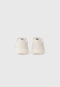 Hvite semskede sneakers med en teksturert overflate, som har en tykk såle og en bakre flik for enkel påkledning. Merking synlig på hælen.