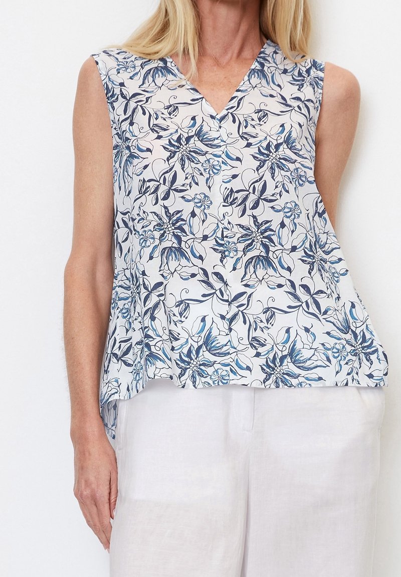 Top senza maniche con motivo floreale blu e bianco, design a scollo a V e tessuto fluido, abbinato a pantaloni di colore chiaro.