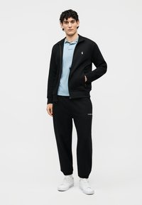 Jaunas vyras stovi vilkėdamas juodą striukę su užtrauktuku, šviesiai mėlyną polo marškinėlį, juodas sportines kelnes ir baltus sportinius batelius, fone yra vienspalvis baltas fonas.