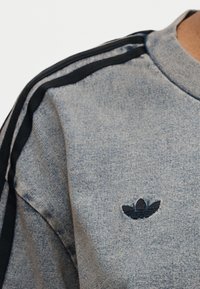 Κοντινό πλάνο ενός γκρι υφασμάτινου πουκαμίσου με το μαύρο λογότυπο trefoil της Adidas στο στήθος και δύο μαύρες ρίγες που κατεβαίνουν στον ώμο.