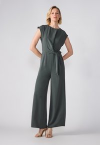 Jumpsuit verde scuro a gamba larga con scollatura avvolgente e cintura a cravatta. Maniche corte e tessuto morbido e leggero. Indossato con tacchi dorati con cinturini.