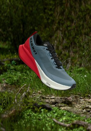 Modro-červená trailová bežecká obuv s označením Mammut, vznášajúca sa nad skalnatým lesným povrchom so zeleným listnatým porastom v pozadí.