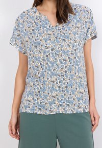 Camisa de botones floral en crema claro, con patrones de flores en azul y marrón, mangas cortas y escote en V. Tejido ligero.