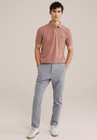 Rozā krāsas polo krekls ar īsām piedurknēm, diskretā logo uz krūtīm, kopā ar gaiši pelēkiem rūtainiem biksēm un baltām kedām.