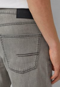 Szare dżinsy z denimu z czarną skórzaną naszywką w pasie, wyraźnym przeszyciem kieszeni i gładkim, teksturowanym wykończeniem.
