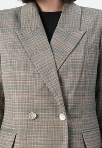 Grå rutete dobbelspent blazer med skarpe revers, teksturert stoff og sølvfargede knapper, med intrikate geometriske mønstre.