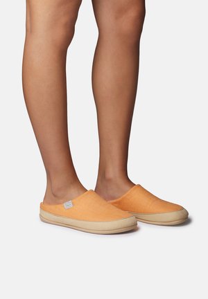 Jambes portant des chaussons orange à enfiler avec des semelles beige, debout devant un fond gris clair uni.