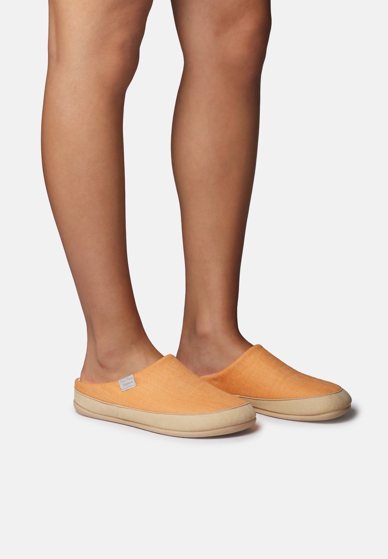 Jambes portant des chaussons orange à enfiler avec des semelles beige, debout devant un fond gris clair uni.
