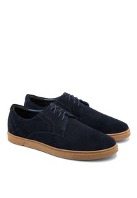Zapatos de ante azul marino con una textura suave, que presentan un diseño clásico de cordones y una suela de goma de color caramelo. Costuras acentuadas en la parte superior.