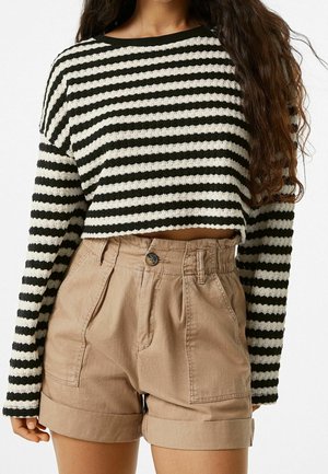Femme portant un pull court rayé noir et blanc à manches longues et un short beige taille haute avec revers, debout devant un fond blanc.