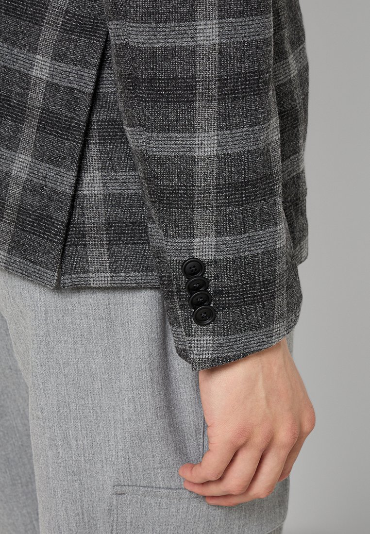 Grau kariertes Blazer mit einem strukturierten Stoff, ausgestattet mit schwarzen Knöpfen an den Manschetten und einer taillierten Silhouette. Tragen über hellgrauen Hosen.