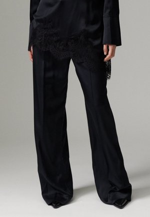 Femme portant un pantalon large noir et un haut noir avec des détails en dentelle, debout devant un fond clair uni.