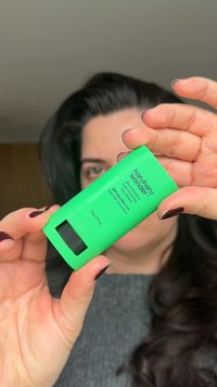 Grön cylindrisk förpackning för solskyddskräm med svart och vit text. Innehåller SPF 50+, storlek 20g. Texturen verkar slät och matt.