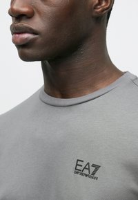 Pelēka t-krekls ar apaļu kakla izgriezumu, izgatavots no mīksta auduma, ar mazu melnu EA7 Emporio Armani logotipu kreisajā krūtīs. Gluda tekstūra, vienkāršs dizains.