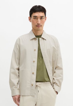 Mężczyzna ubrany w beżową zapinaną na guziki kurtkę na oliwkowej zielonej koszuli i beżowe spodnie ze sznurkiem, stojący na białym tle.
