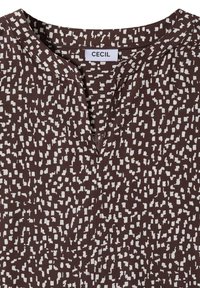 Chemise marron avec motif carré abstrait blanc, col rond avec une petite ouverture à l'avant, et une étiquette indiquant « CECIL » à l'intérieur du col.