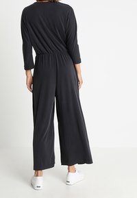 Jumpsuit noir à manches longues avec une taille élastique et des jambes larges, fabriqué dans un tissu doux. Associé à des baskets blanches pour le contraste.