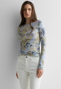 Mujer con una camiseta ajustada de manga larga con patrones florales en beige y azul y pantalones blancos, de pie frente a un fondo liso de color gris claro.
