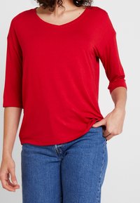 Top rosso in tessuto morbido con scollatura a V e maniche a tre quarti, vestibilità comoda, texture liscia, abbinato a jeans blu. Nessun motivo o accento visibile.