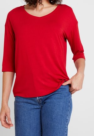 Top rosso in tessuto morbido con scollatura a V e maniche a tre quarti, vestibilità comoda, texture liscia, abbinato a jeans blu. Nessun motivo o accento visibile.