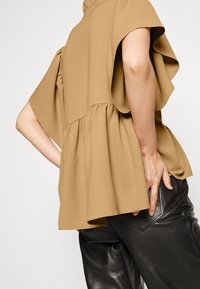 Top à manches courtes avec basque en tissu beige, doté d'une coupe ample, d'une surface texturée et d'accents à volants aux épaules. Associé à un pantalon noir.