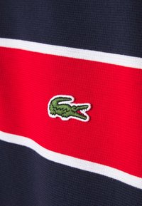 Logo crocodile vert brodé sur un tissu tricoté rayé rouge et bleu marine avec de fines bordures blanches.