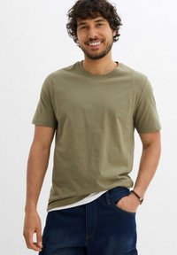 Uomo sorridente con capelli ricci, indossa una maglietta verde oliva sopra una camicia bianca e jeans blu, con una mano in tasca, in piedi davanti a uno sfondo uniforme.