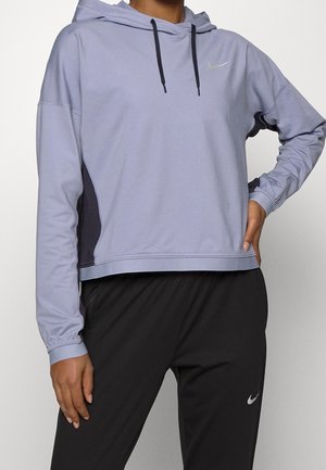 Persoon die een lichtpaarse Nike hoodie draagt met zwarte trekkoorden en zwarte Nike sportbroek, poseert met één hand op de heup.