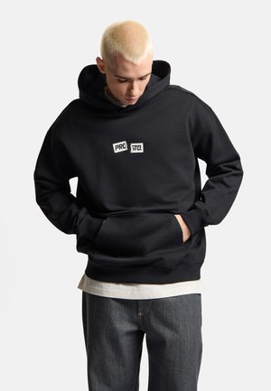 Młoda osoba z krótkimi blond włosami, ubrana w czarną bluzę z kapturem z napisem "PROSTO.", ręce w przedniej kieszeni, spogląda w dół.