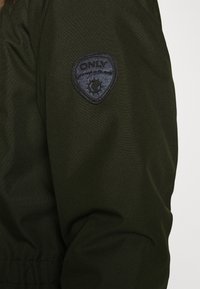 Material de chaqueta verde oliva con una textura suave, que presenta un parche de logo bordado negro "ONLY" en la manga. Detalle de dobladillo elástico.
