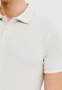 Jasnoszara koszulka polo wykonana z teksturowanej tkaniny. Posiada klasyczny kołnierzyk, zapięcie na dwa guziki oraz subtelnie haftowane logo na piersi.