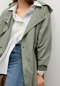 Lichtgroene, waterafstotende trenchcoat met capuchon, voorzien van een losse pasvorm, subtiele glans en een zwart trekkoord. Gelaagd over een wit-blauw gestreept overhemd.