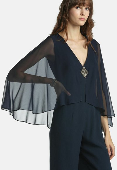 Marineblaue, durchsichtige Bluse mit tiefem V-Ausschnitt und weiten Umhangärmeln, vorne verziert mit einer geometrischen Brosche, kombiniert mit dunkler Hose.