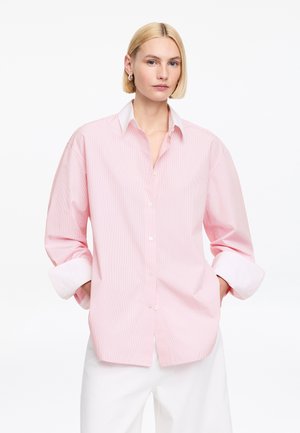 Blondína s krátkymi vlasmi, oblečená v nadmernej ružovej a bielej pruhovanej košeli s vyhrnutými rukávmi a v bielych nohaviciach, stojaca s rukami v vreckách.