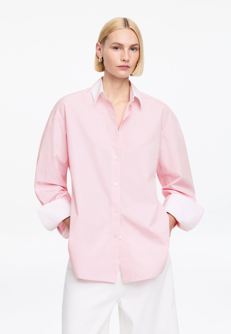 Mujer rubia con cabello corto, vistiendo una camisa de rayas rosa y blanca de gran tamaño con mangas remangadas y pantalones blancos, de pie con las manos en los bolsillos.