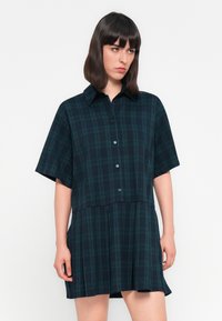 Frau mit dunklem Haar, die ein kurzärmliges, dunkelgrünes und marineblau karierter, geknöpfter Dress trägt, steht vor einem schlichten weißen Hintergrund.