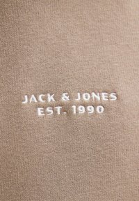 Bežna pamučna tkanina s teksturiranim pletenjem. Bijeli izvezeni tekst glasi "JACK & JONES EST. 1990." Jednostavan, čist dizajn bez dodatnih detalja.