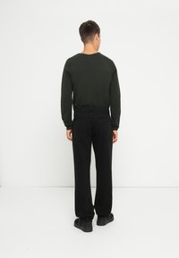 Camicia verde scuro a maniche lunghe abbinata a pantaloni neri a gamba dritta e sneakers neri, vista da dietro su uno sfondo bianco.