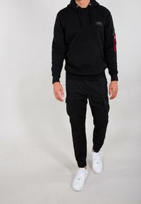 Uomo che indossa una felpa nera con cappuccio e pantaloni cargo con sneakers bianche, che cammina su uno sfondo chiaro e uniforme.
