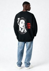 Felpa nera oversize con una grafica raffigurante una donna con una fascia per capelli, accompagnata dal testo "OOPS I DON'T CARE" in rosso. Pantaloni casual di denim blu.