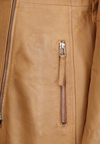 Tan leren jack met een gladde textuur, voorzien van een zichtbare ritszak en metalen ritsbeslag. Stijlvolle stikdetails.