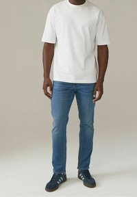 Hombre vestido con una camiseta de manga corta blanca lisa, jeans ajustados azul y zapatillas oscuros con franjas blancas, de pie sobre un fondo claro.