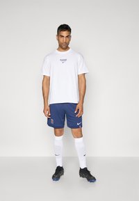 Hvid t-shirt med "PARIS" grafik, marineblå shorts med holdlogo, hvide sokker og sorte fodboldstøvler. Enkel, atletisk design.