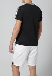 Sort atletisk T-shirt med grå accenter på skuldrene, parret med hvide shorts. T-shirten har en rund halsudskæring og sideslids for bedre bevægelsesfrihed.