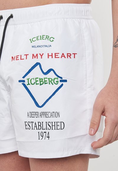 Costumi da bagno bianchi con vita regolabile con cordino, grafica stampata in rosso, blu e verde, con elementi di branding e testo. Materiale leggero.